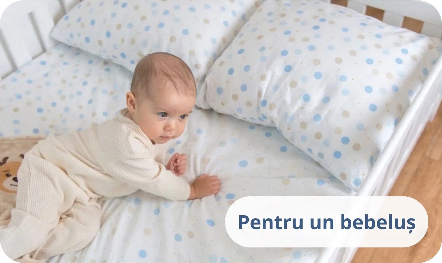 Bebeluș stând pe burtă pe un pat alb în lenjerie cu buline albastre.